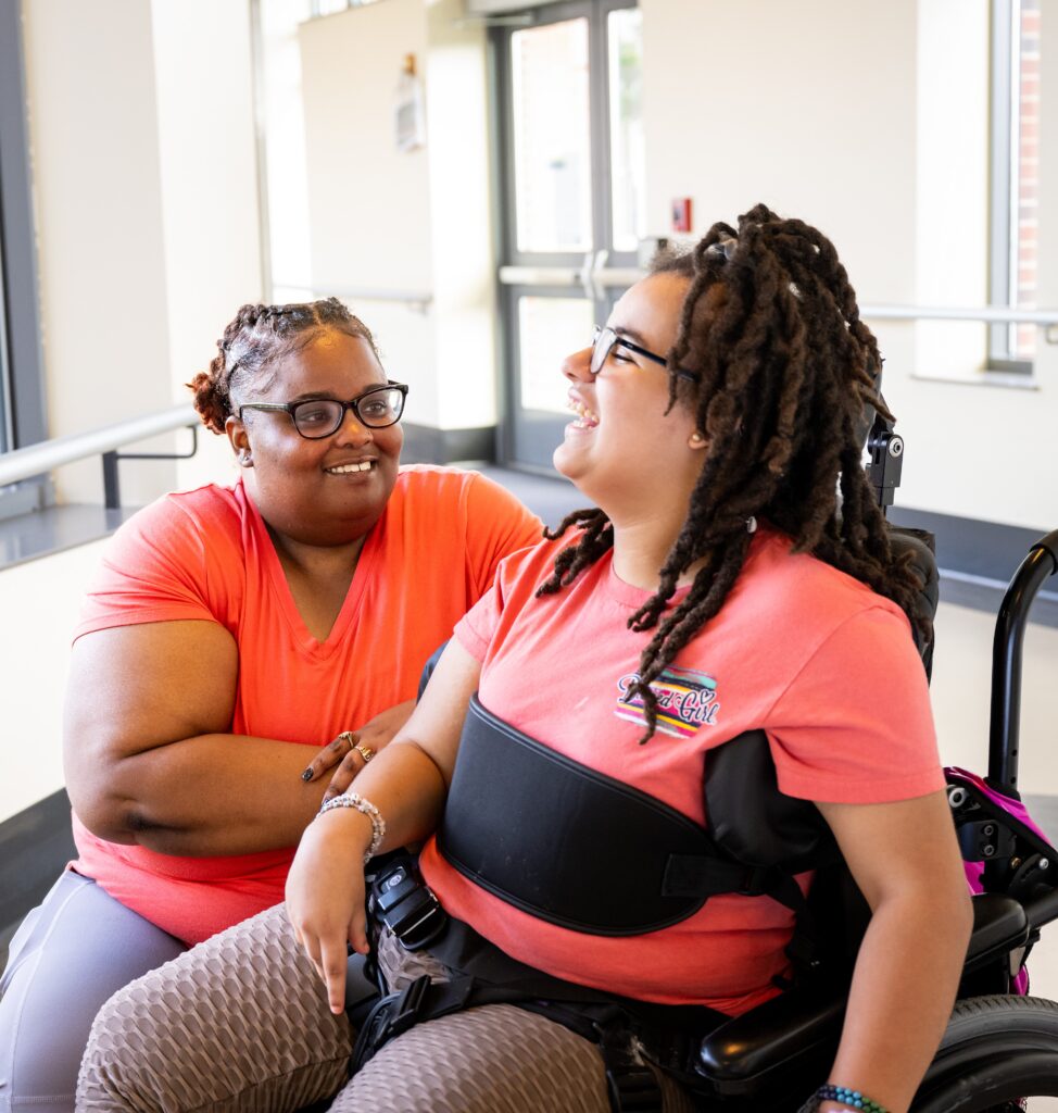 Two people, both wearing orange shirts, share a moment of companionship. One is kneeling while the other is sitting in a wheelchair. They are smiling and interacting in a brightly lit room.