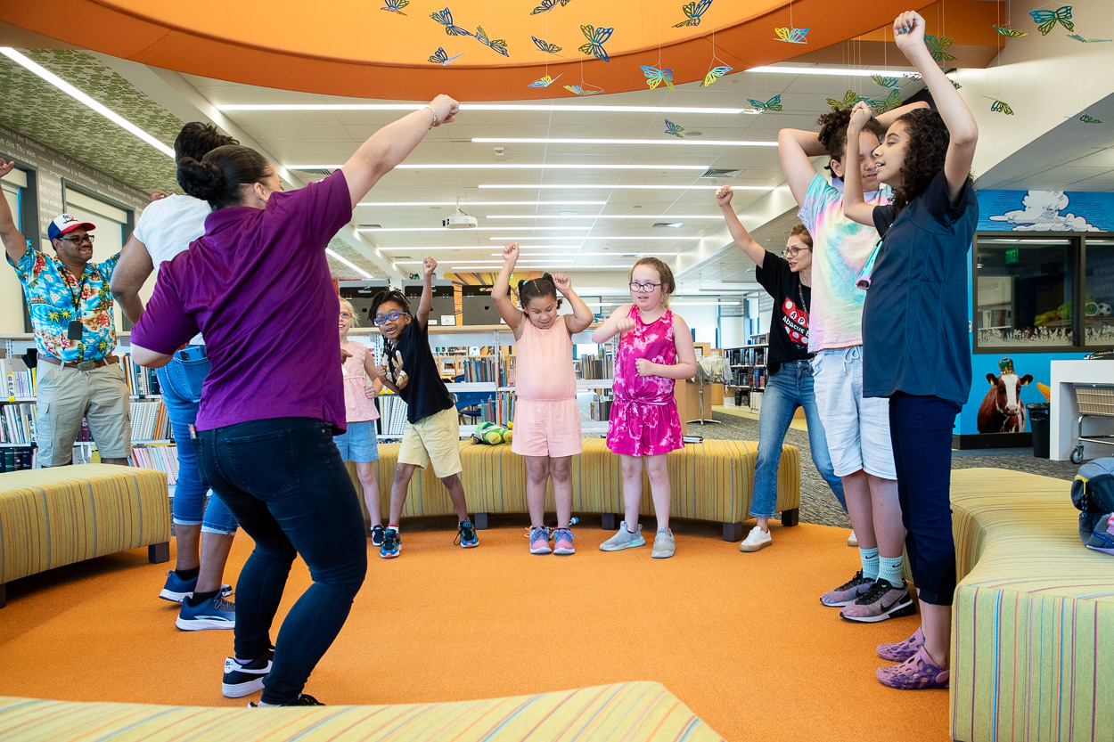 A group of children and adults participate in an activity inside a library, standing in a circle with raised arms.