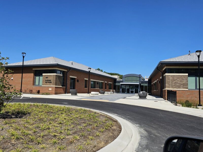 A modern brick building with a curved driveway and landscaped area in the forefront, under a clear blue sky. The entrance is flanked by two single-story structures with large windows.